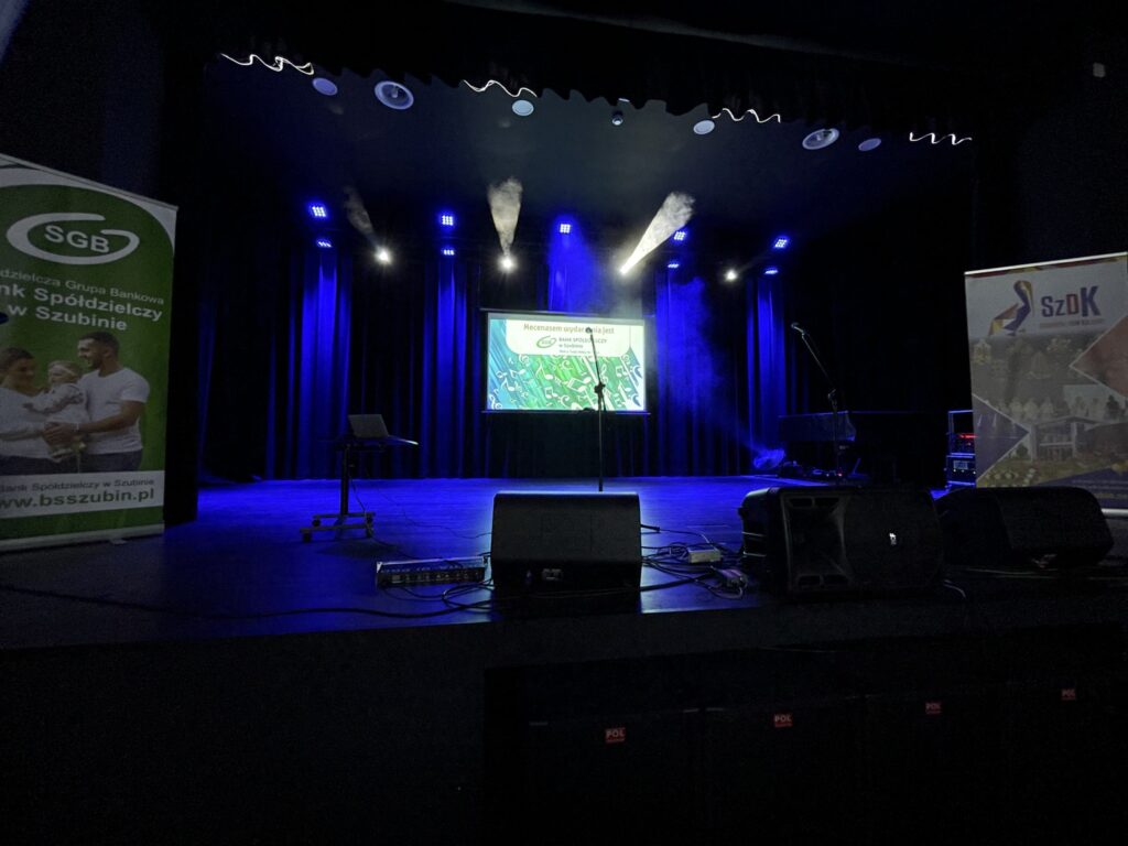 Wnętrze sali teatralnej z oświetleniem, pianinem i ekranem prezentacyjnym SGB oraz SzDK.