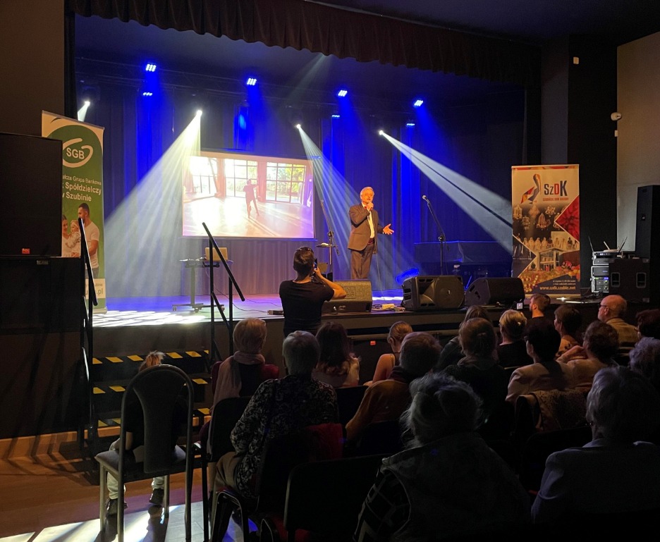 Wystąpienie na scenie w sali z oświetleniem, publiczność wpatrzona w prelegenta na konferencji.