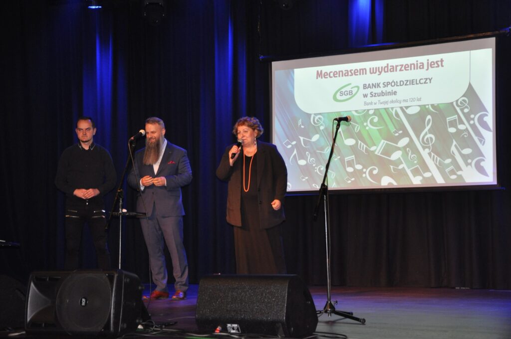 Uczestnicy wydarzenia artystycznego na scenie, z projekcją muzyczną w tle. Sponsorem jest Bank Spółdzielczy.