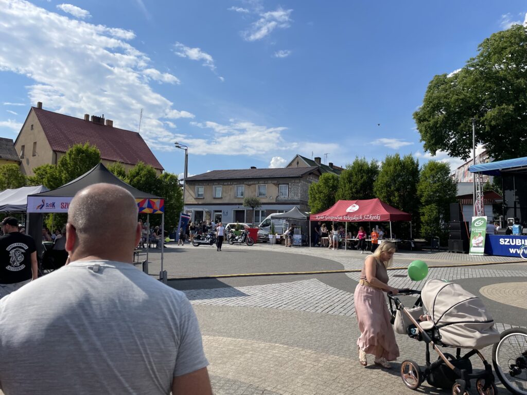Tłum ludzi na festynie w mieście, stragany, dzieci z balonami i kobieta z wózkiem spacerowym w słońcu.