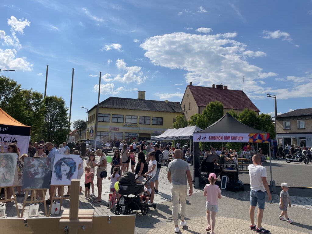 Tłum festiwalu w Szubinie, z ustawionymi stoiskami i artystami na placu w słoneczny dzień.