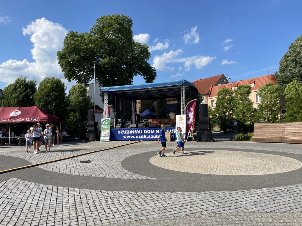 Tłum ludzi na placu podczas wydarzenia kulturalnego, z nową sceną i zielonymi drzewami w tle.