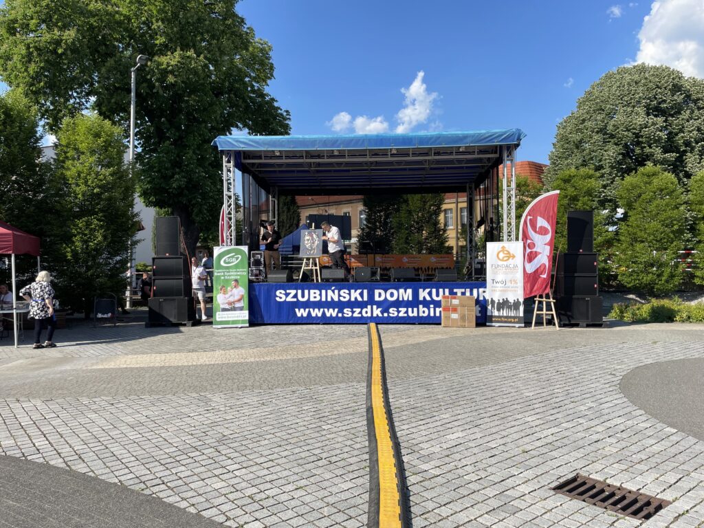 Scena plenerowa z logo Szubińskiego Domu Kultury, otoczona zielenią i festiwalowymi stoiskami.