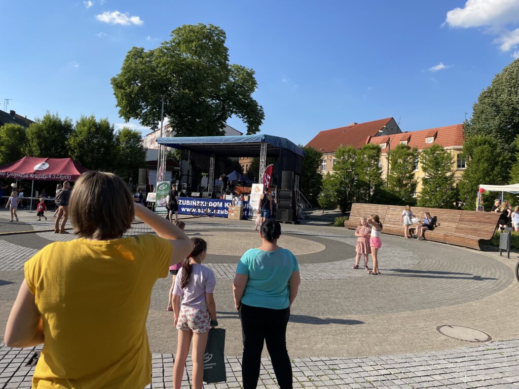 Ludzie bawią się na festiwalu w parku, z sceną i straganami w tle.