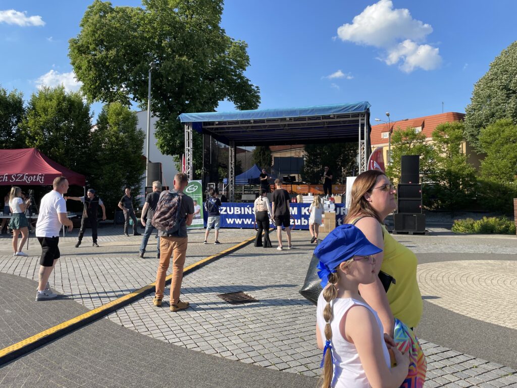 Ludzie uczestniczą w wydarzeniu plenerowym, obok sceny i stoisk, w słoneczny dzień.