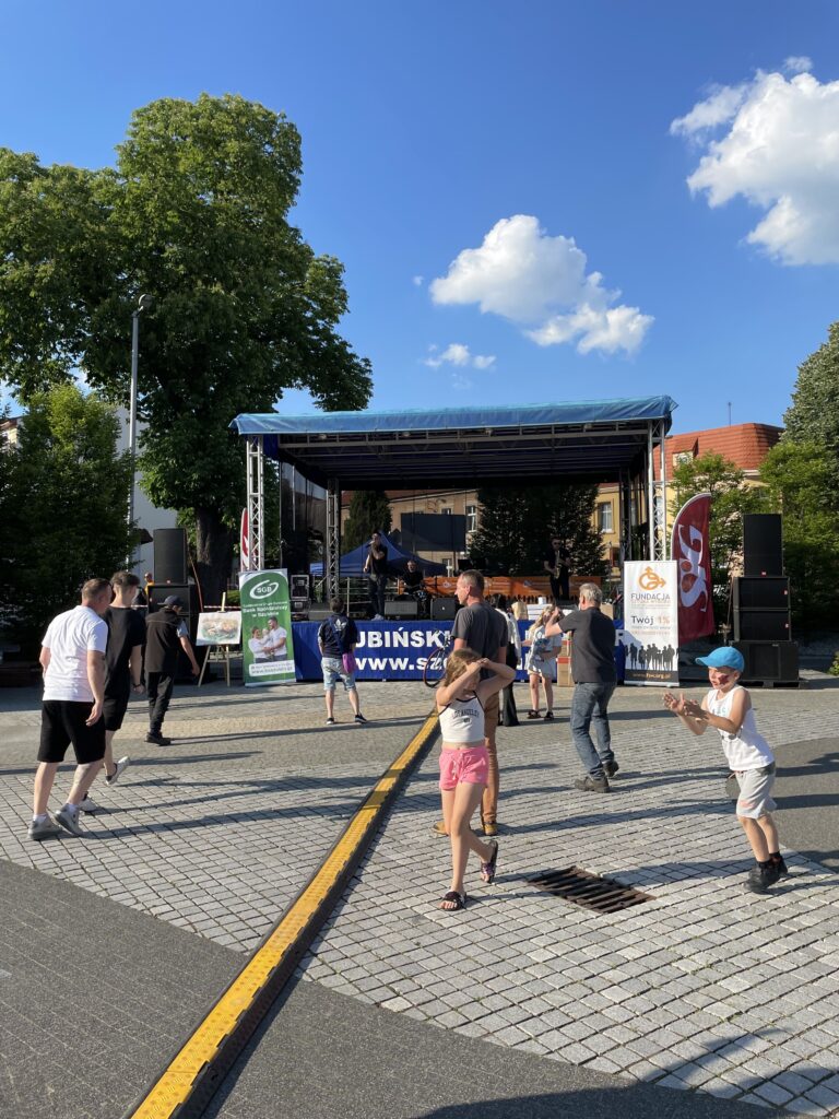 Uczestnicy festiwalu tańczą przed sceną, w słoneczny dzień, w otoczeniu zieleni i niebieskiego nieba.