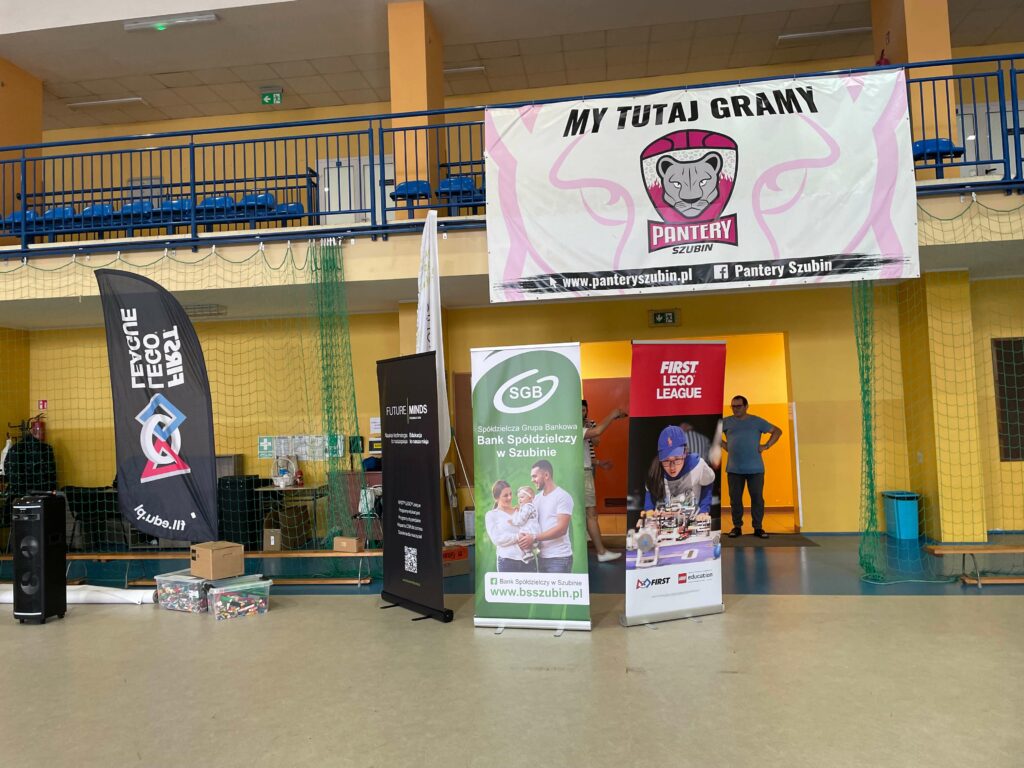 Widok hali sportowej z banerami, w tym "Pierwsza Liga LEGO" i "Pantery Szubin" oraz stoiska sponsorów.