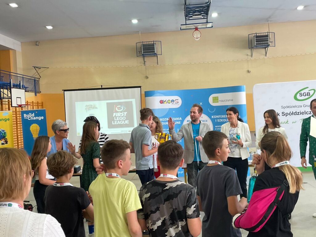 Uczestnicy wydarzenia związanego z First Lego League w hali sportowej, z prelegentami i uczestnikami.